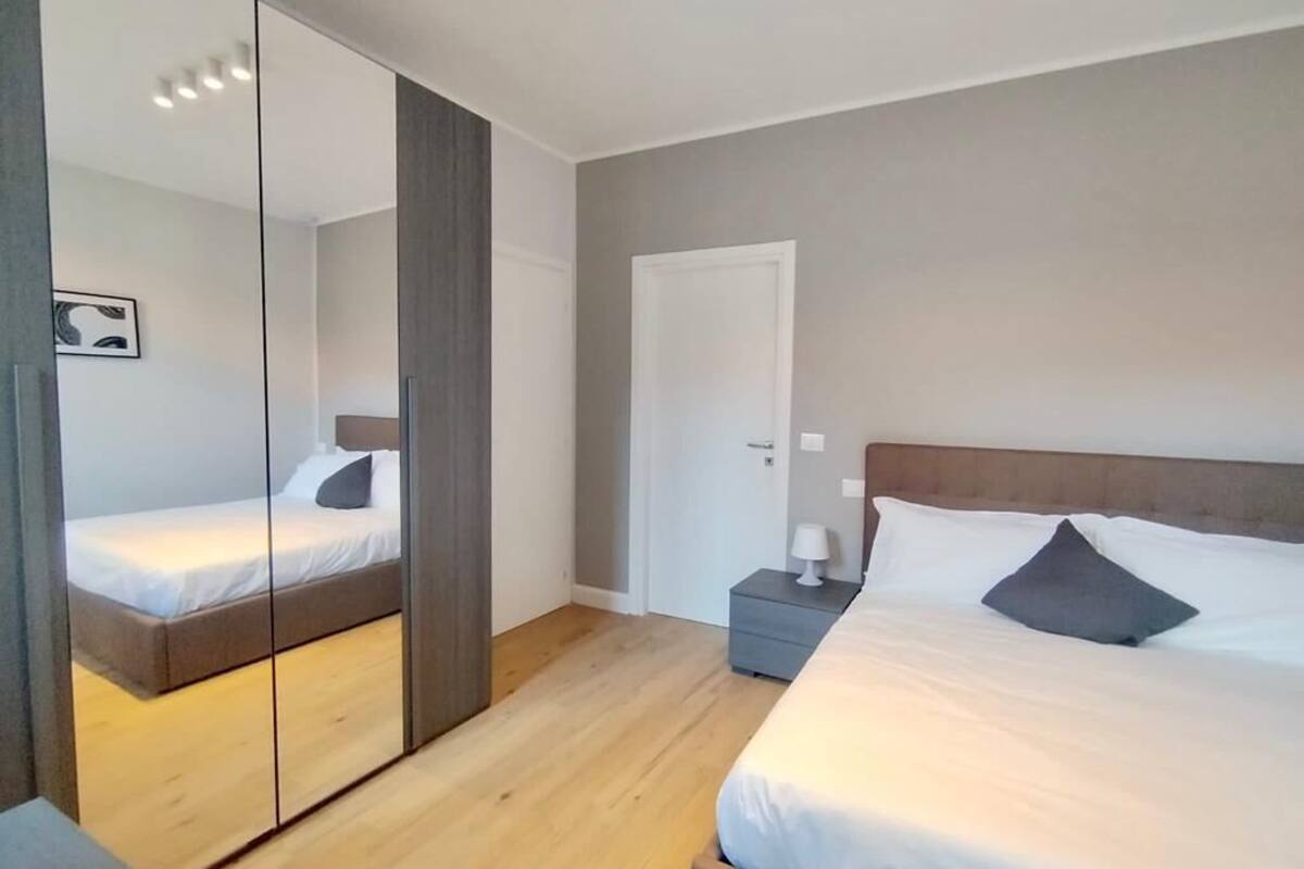 A modern bedroom with a large mirror, a wooden floor, a bed with white bedding, a nightstand with a lamp, and a door.
