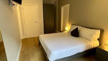Un dormitorio con cama, mesitas de noche, televisor montado en la pared y una puerta que da a otra habitación.