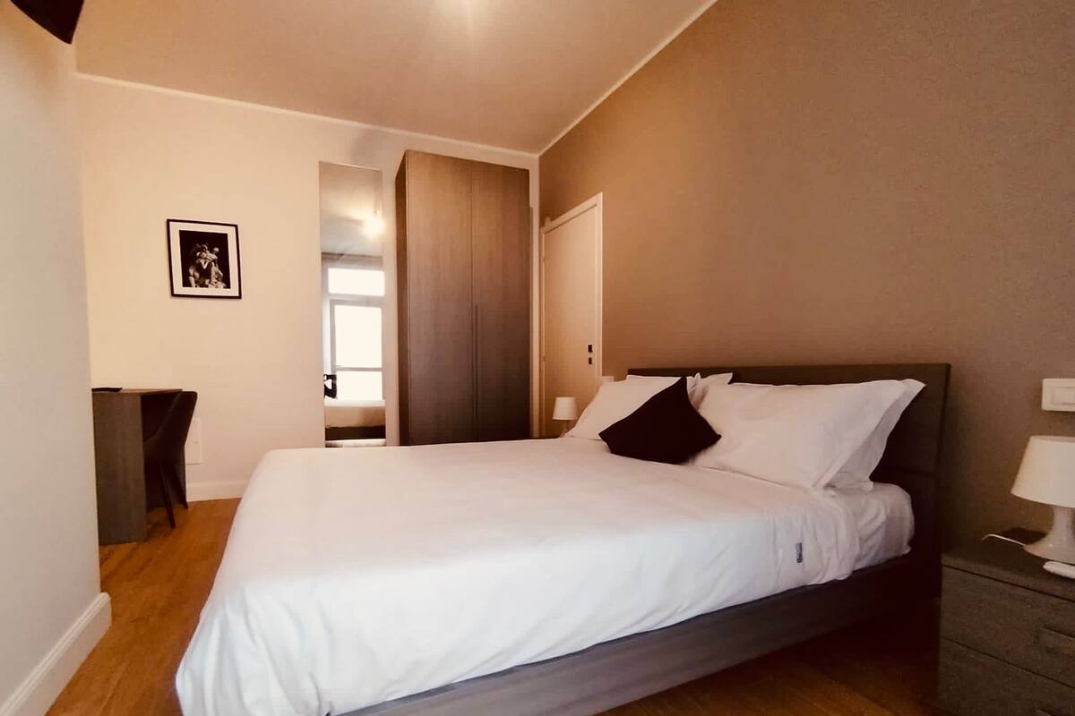 A neatly made bed with white linens and a dark headboard in a room with wooden flooring and a door leading to another room.