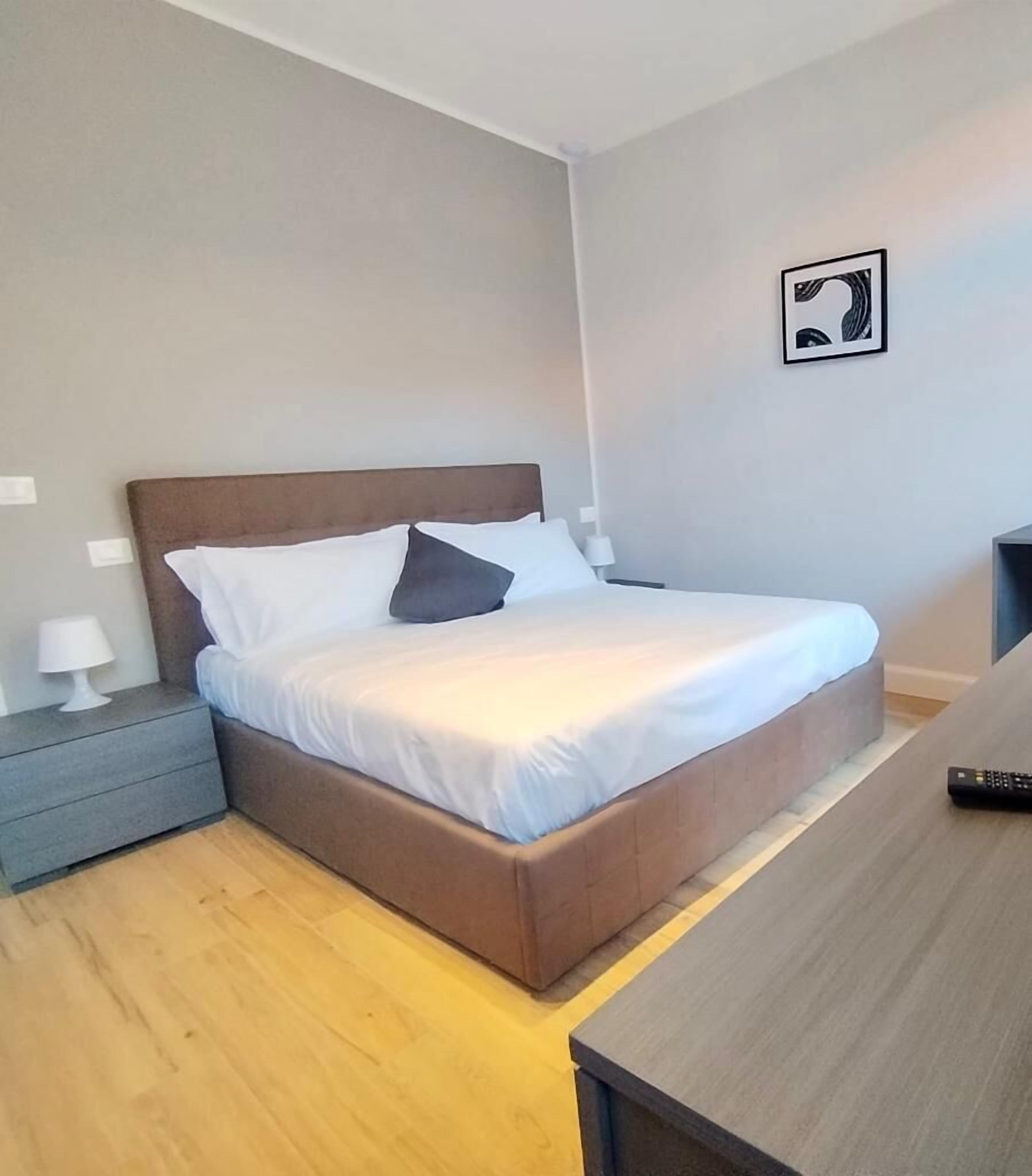 Une chambre à coucher avec un lit, des tables de chevet, une commode, une lampe, un tableau encadré au mur et une télécommande posée sur une table.