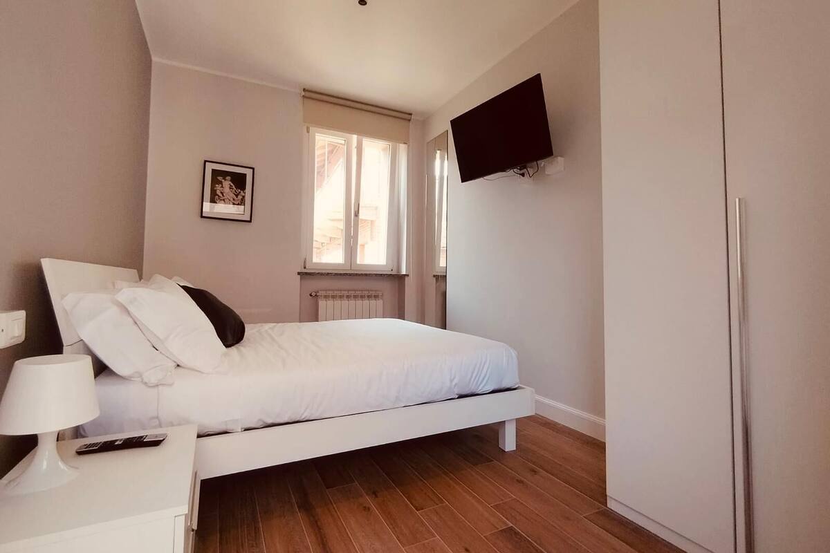 A bedroom with a bed, a bedside table with a lamp, a TV mounted on the wall, and a window with a view of buildings.
