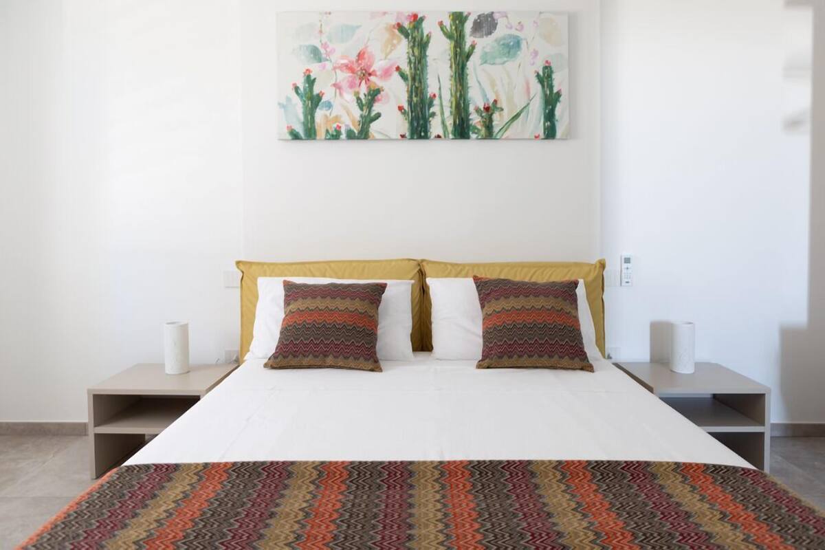 A neatly made bed with a patterned bedspread, two pillows, and a wall-mounted painting of cacti and flowers.