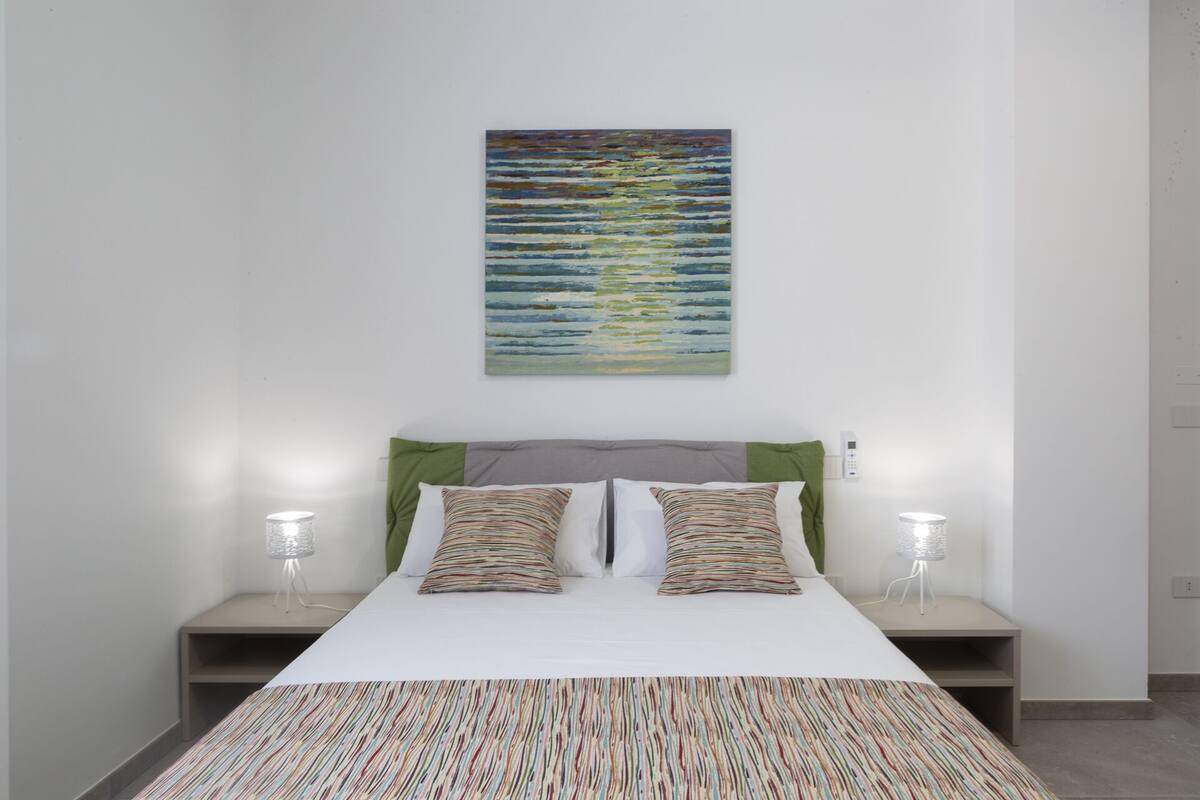 A modern bedroom with a bed, two bedside tables, a nightstand, a wall-mounted painting, and a floor lamp.