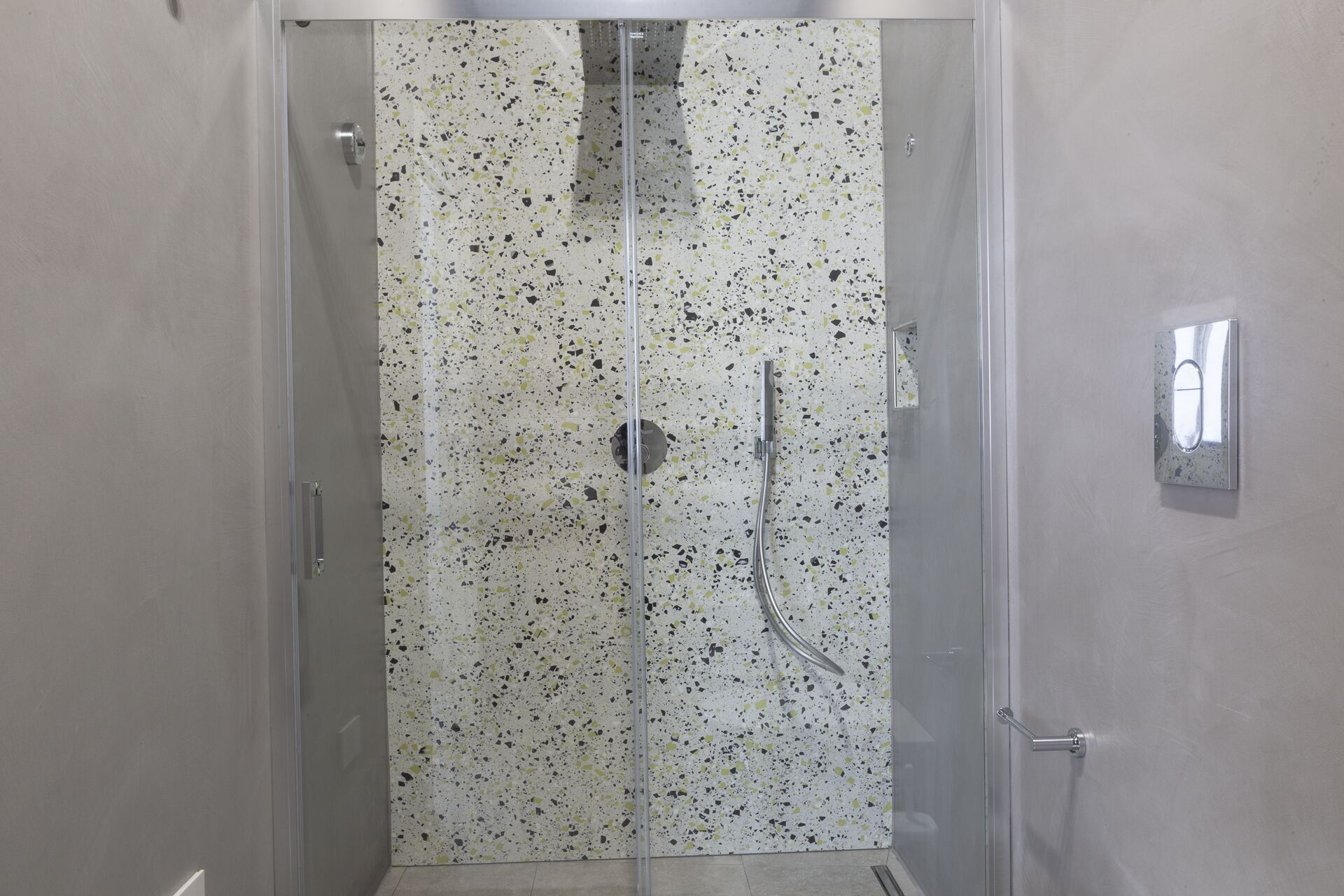 A glass-enclosed shower with a speckled tile wall, a handheld showerhead, and a wall-mounted faucet.