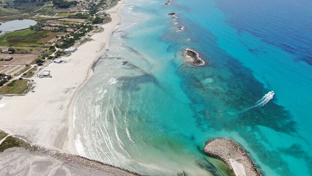 Vista aérea de una zona costera con playa, aguas azules cristalinas y una combinación de terreno natural y urbanizado.