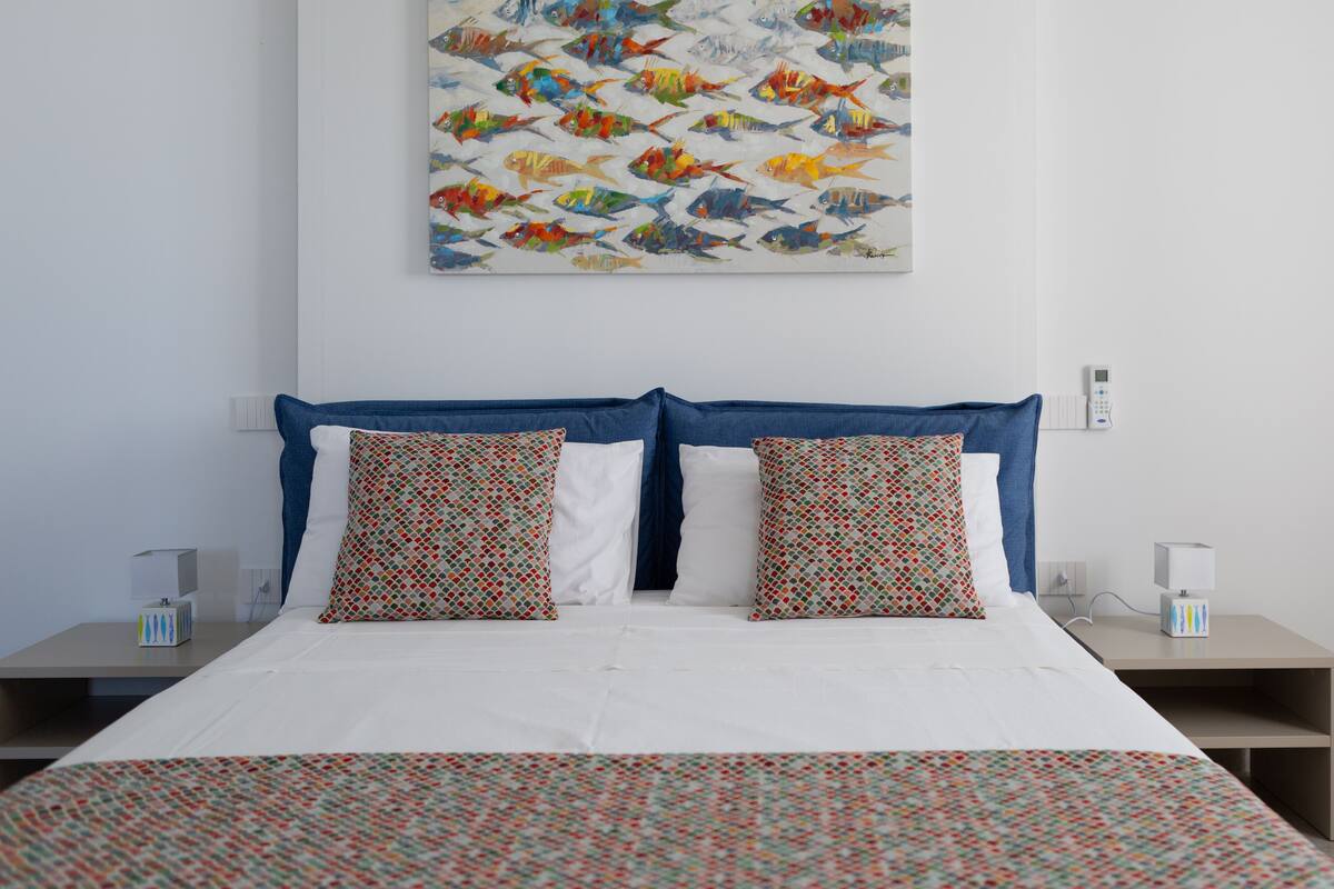 A neatly made bed with patterned bedding, a wall-mounted artwork of colorful fish, and bedside tables with lamps and tissue boxes.