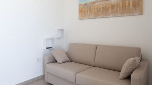 Un sofá beige con dos cojines, una pared blanca y un cuadro de un paisaje.