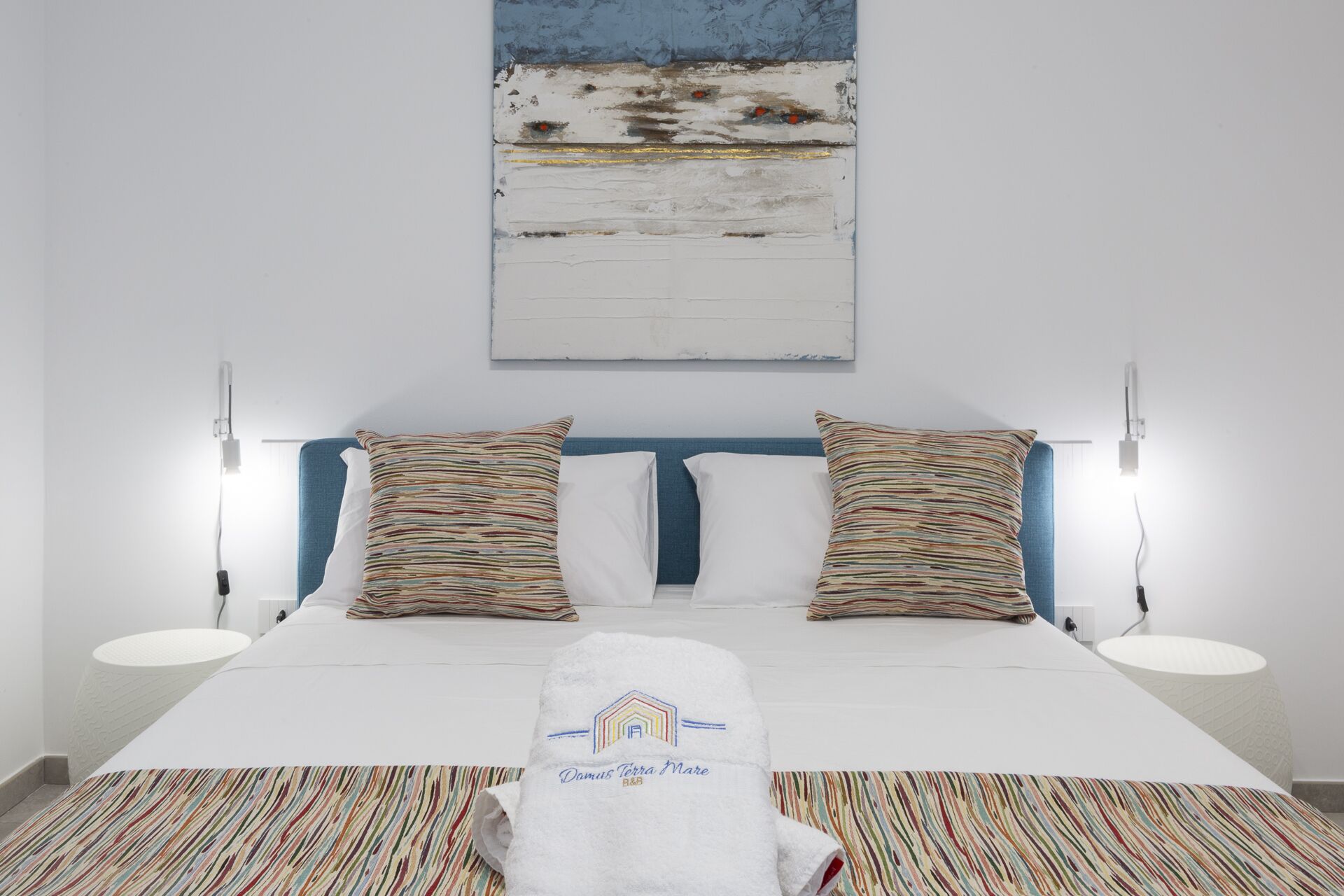 A neatly made bed with striped pillows, a white towel with a logo, and a painting above it.