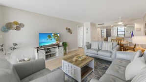 A modern living room with a flat-screen TV, grey sofas, a coffee table, and a kitchen in the background. - SW-Facing Beachfront Condo | Pool/Gym/Pickleball (Marco Island)
