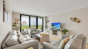A modern living room with a sofa set, a coffee table, a TV, and a balcony with a view. - SW-Facing Beachfront Condo | Pool/Gym/Pickleball (Marco Island)