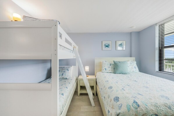 A bedroom with bunk beds, a bedside table with a lamp, a bed with a patterned bedspread, and two framed pictures on the wall.