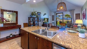 A kitchen with granite countertops, stainless steel sinks, and wooden cabinets. There is a dining area with a view of palm trees and a pool.