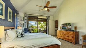 A bedroom with a bed, a ceiling fan, a TV, a wooden dresser, and a balcony with a view of palm trees.