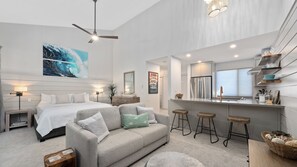 A modern bedroom with a large bed, a sitting area with a sofa, a ceiling fan, and a kitchen area with a refrigerator and bar stools.