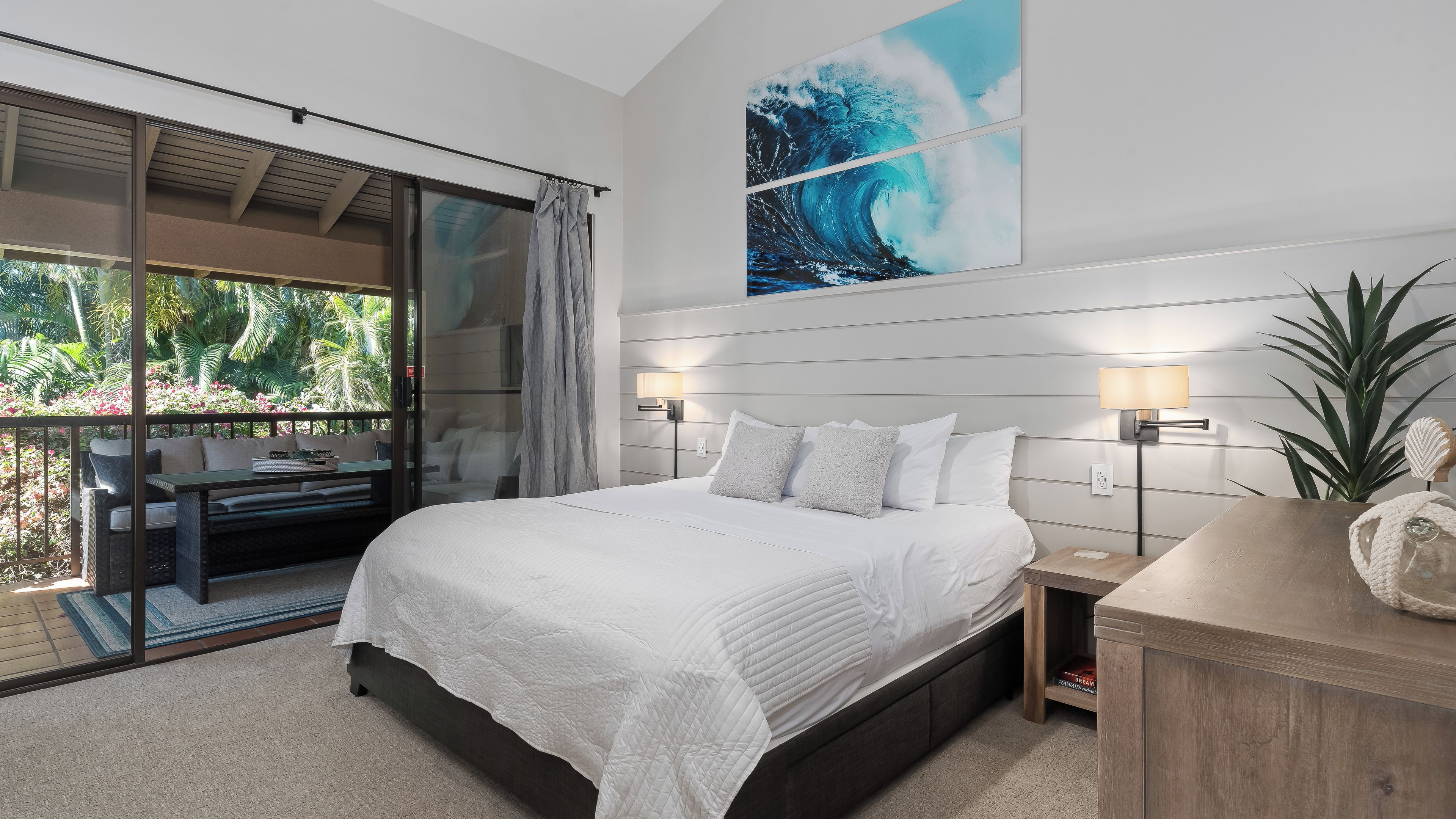 A bedroom with a large bed, a nightstand, a wall-mounted lamp, and a view of a balcony with outdoor furniture.