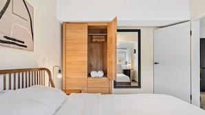A bedroom with a wooden wardrobe, a bed with white linens, and a mirror reflecting a bathroom.