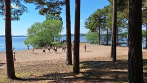 Uma praia com pessoas jogando vôlei, cercada por árvores altas e um lago ao fundo.