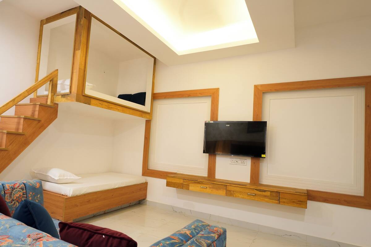 A room with a wooden staircase, a bed with a headboard, a TV mounted on the wall, and a small table with drawers.