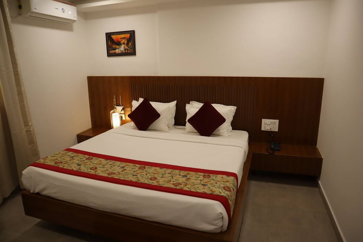 A neatly made bed with a floral bedspread and two decorative pillows, a bedside table with a lamp, and a wall-mounted air conditioner.
