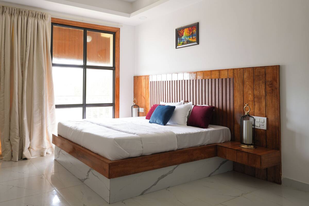A bedroom with a wooden headboard, a bed with white bedding, a nightstand with a lamp, a window with curtains, and a painting on the wall.