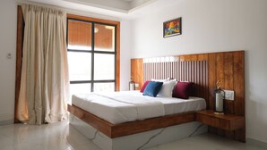 A bedroom with a wooden headboard, a bed with white bedding, a nightstand with a lamp, a window with curtains, and a painting on the wall.