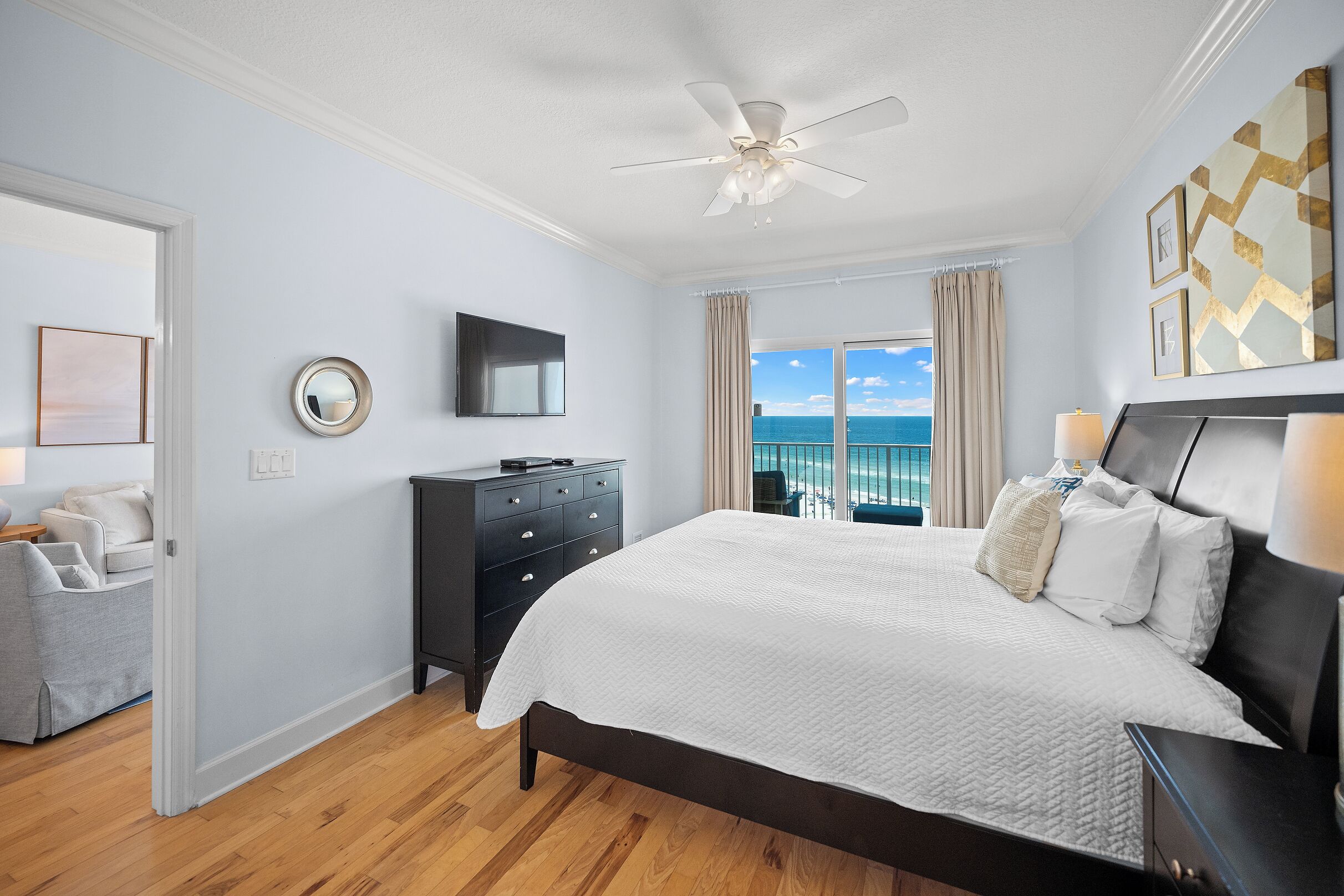 A bedroom with a bed, a ceiling fan, a TV, a dresser, a view of the ocean, and a balcony.