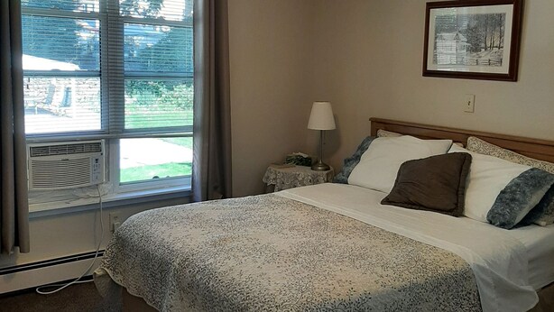 A bedroom with a bed, a ceiling fan, a window with blinds, a bedside table with a lamp, and a framed picture on the wall. - Trillium Bed & Breakfast (Niagara Falls)