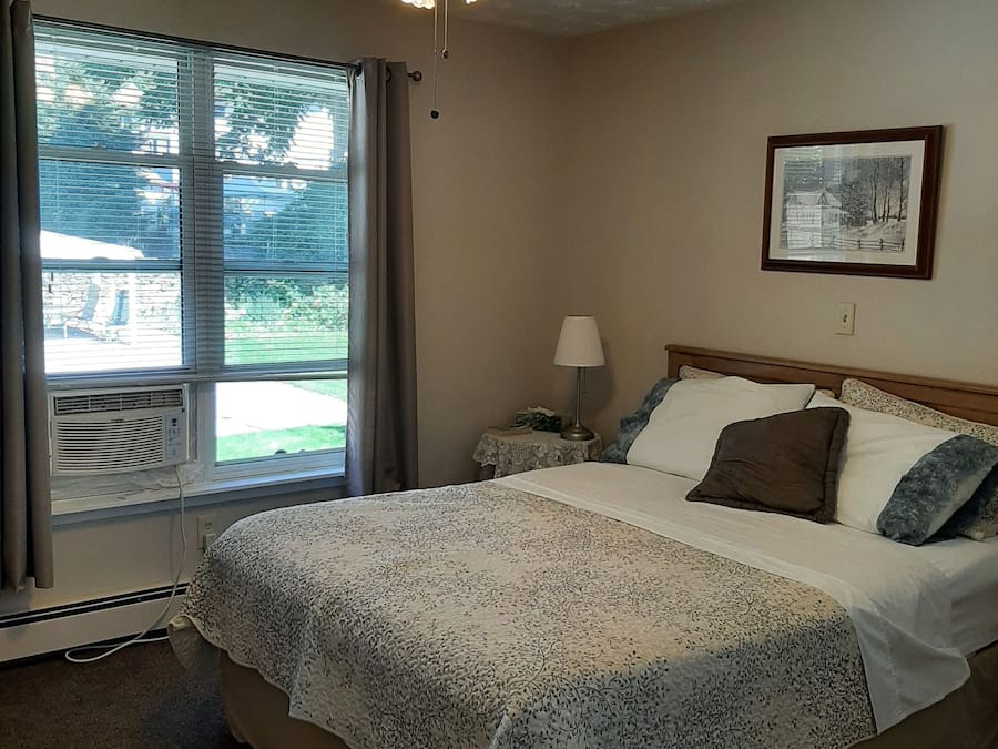 Une chambre à coucher avec un lit, un ventilateur de plafond, une fenêtre avec des stores, une table de chevet avec une lampe et un tableau encadré au mur.