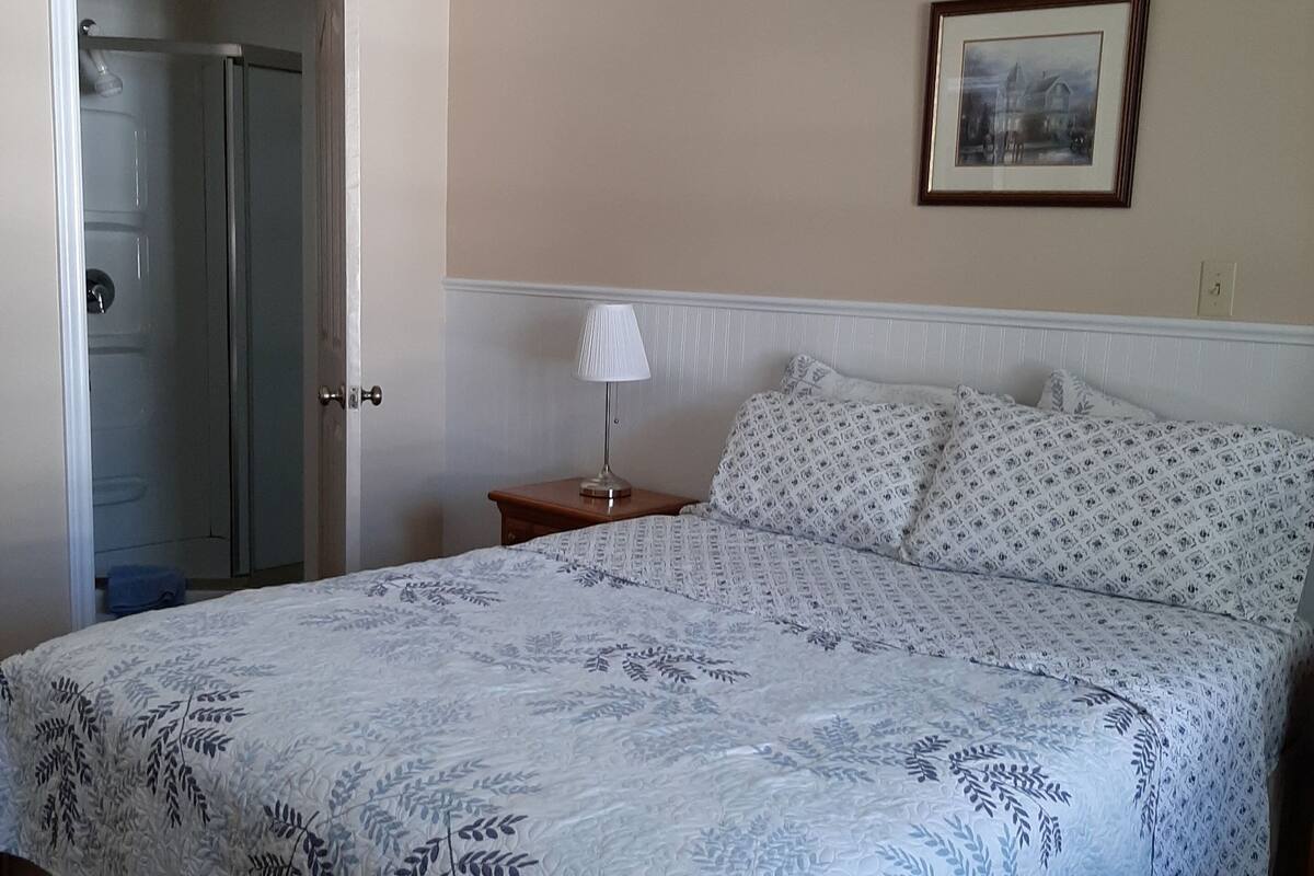 A bedroom with a bed, a nightstand, a lamp, a framed picture on the wall, and a door leading to a bathroom.