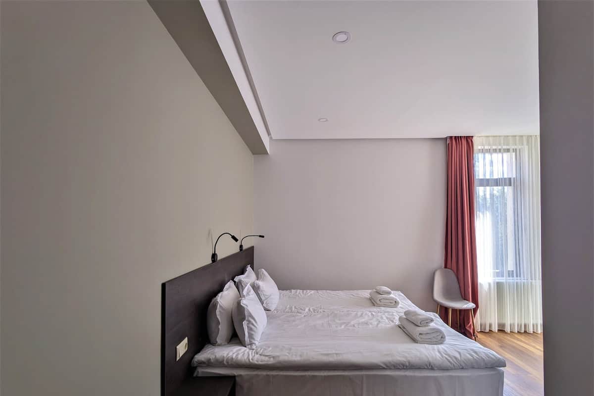 A neatly made bed with pillows and a rolled towel, a bedside table, a chair, and a window with curtains.