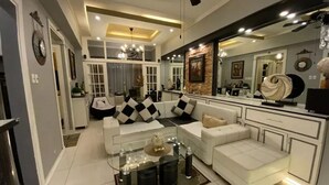 Een moderne woonkamer met een glazen salontafel, witte banken en een keukenhoek met een grote spiegel en aanrecht.