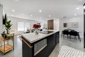 Een moderne keuken met een centraal eiland, een eetgedeelte met een zwarte tafel en stoelen, en een zithoek met een bank en salontafel.