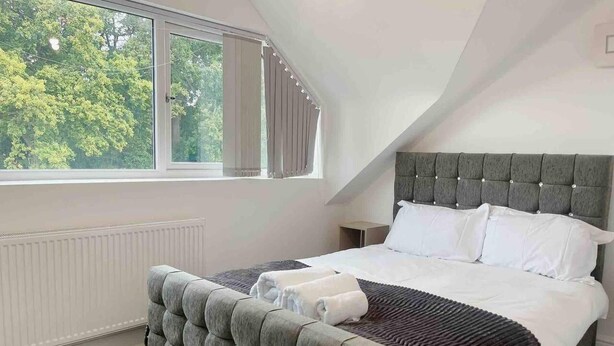 Ein Schlafzimmer mit einem großen Bett, einem grau gepolsterten Kopfteil, einem Fenster mit Vorhängen und einer Heizung.