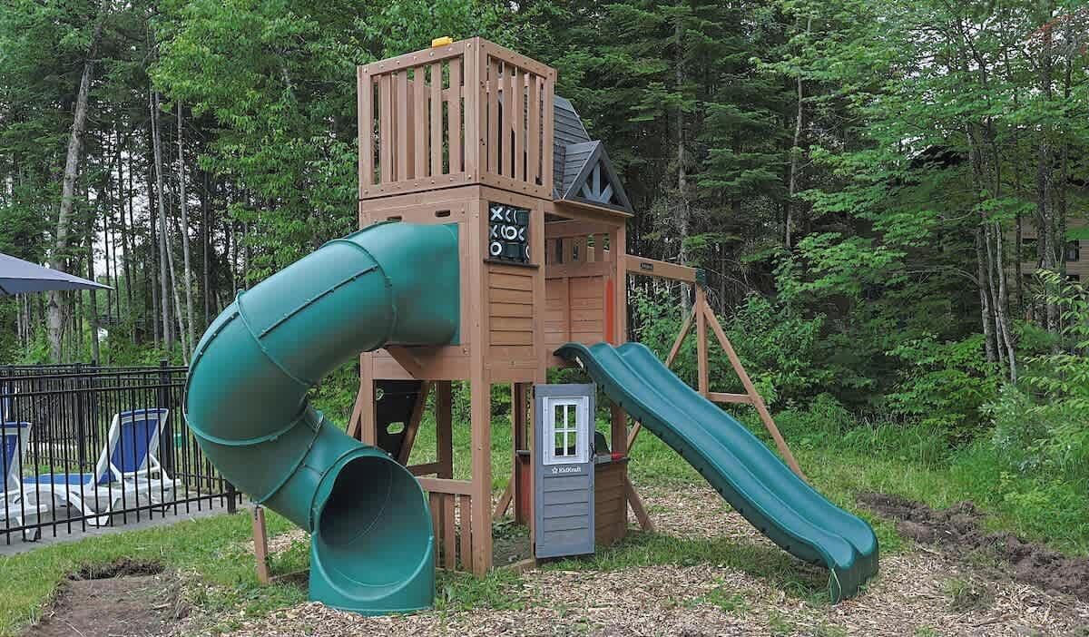 A wooden play structure with a slide and a climbing wall in a wooded area.