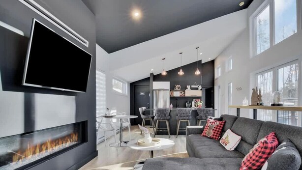 A modern living room with a fireplace, a TV mounted on the wall, a grey sofa with red and black pillows, and a dining area with a bar. - Cerf Blanc | Sauna & Piscine by Alchalet (Pont-Rouge)