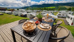 Una terraza de madera con una mesa dispuesta para el desayuno, rodeada de vehículos recreativos en un camping con un lago y colinas al fondo.