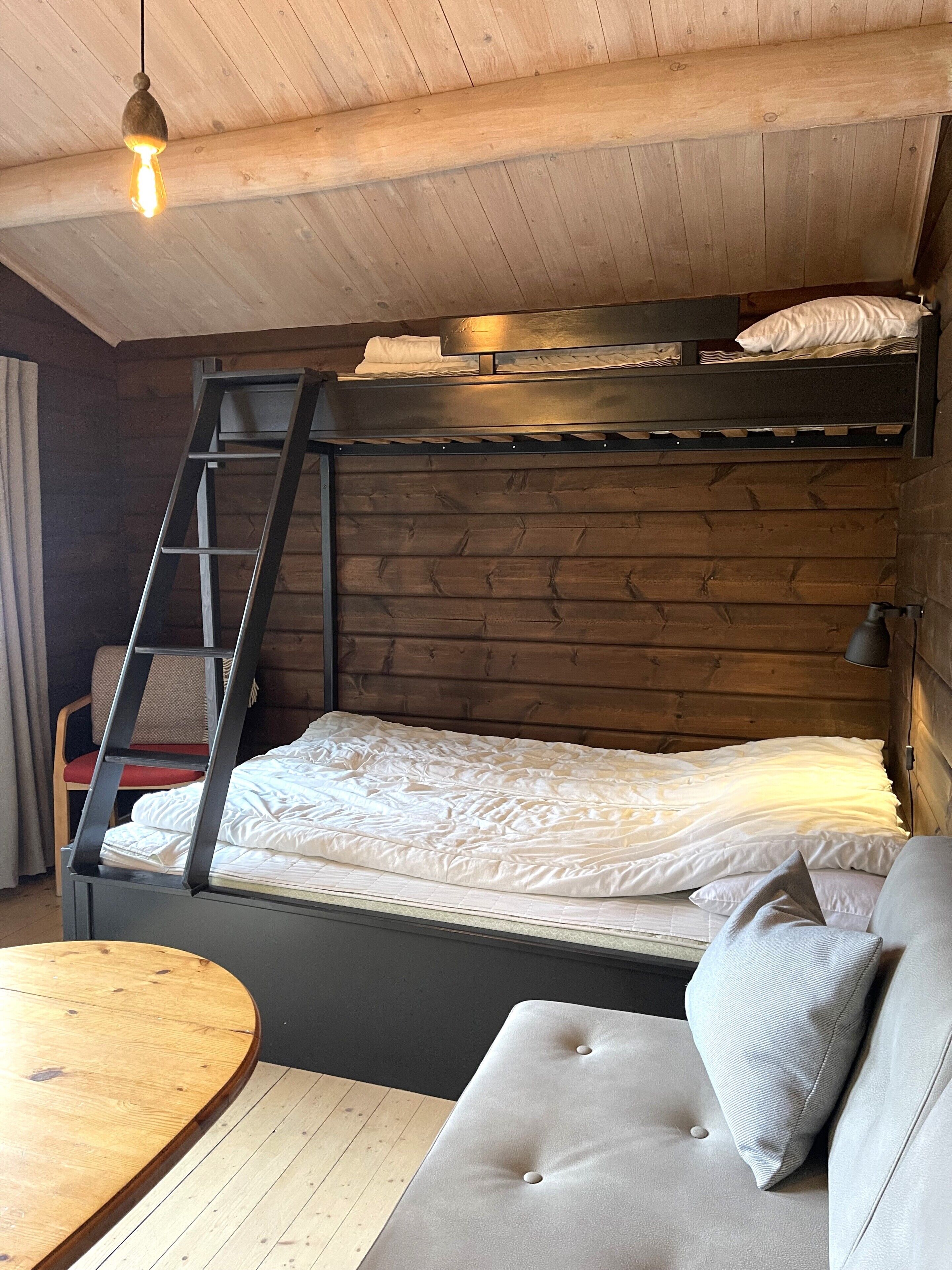 A bunk bed with a ladder, a small table, and a sofa in a room with wooden walls and a wooden ceiling.