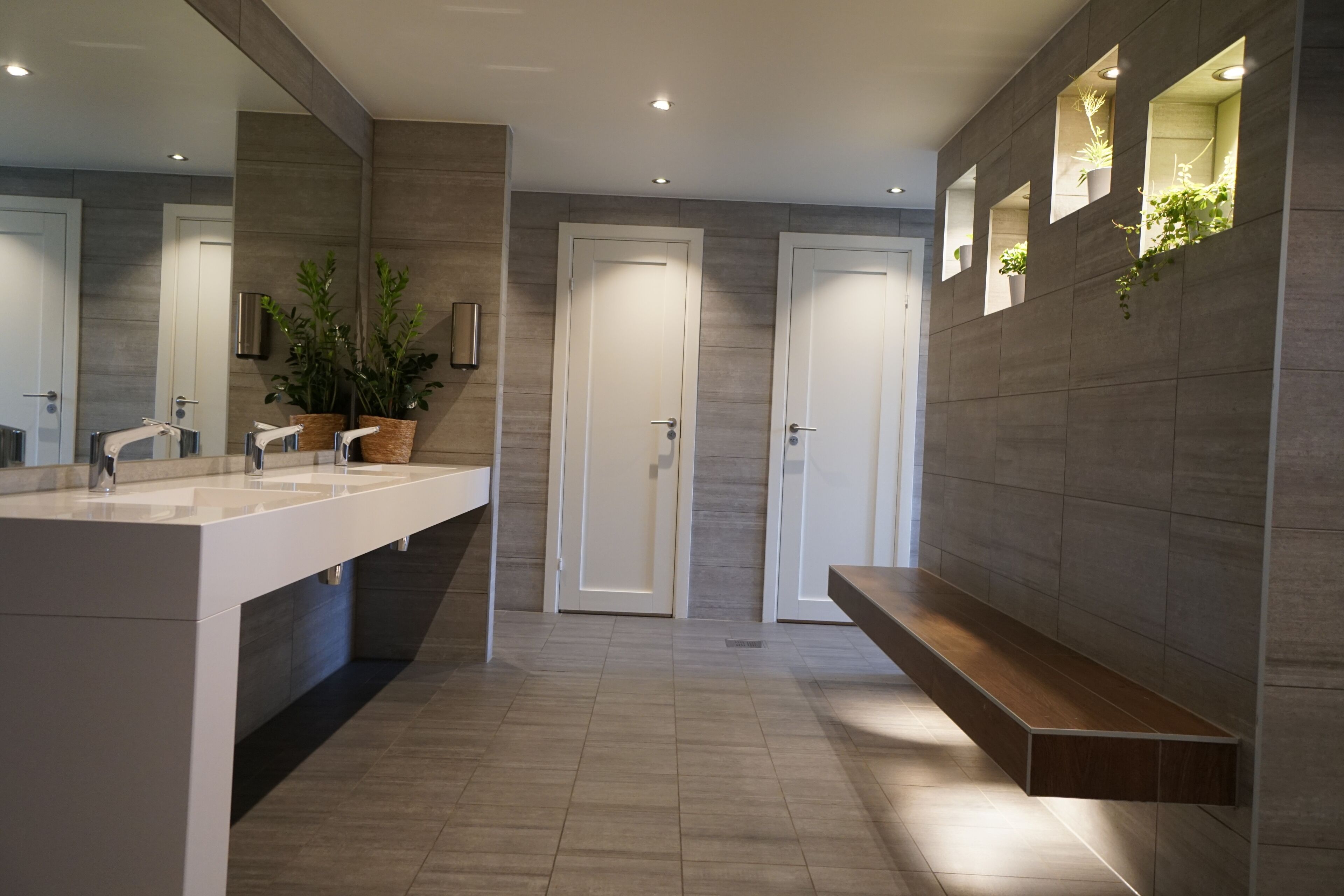 Un baño moderno con un espejo grande, dos lavamanos, un banco y plantas en macetas.