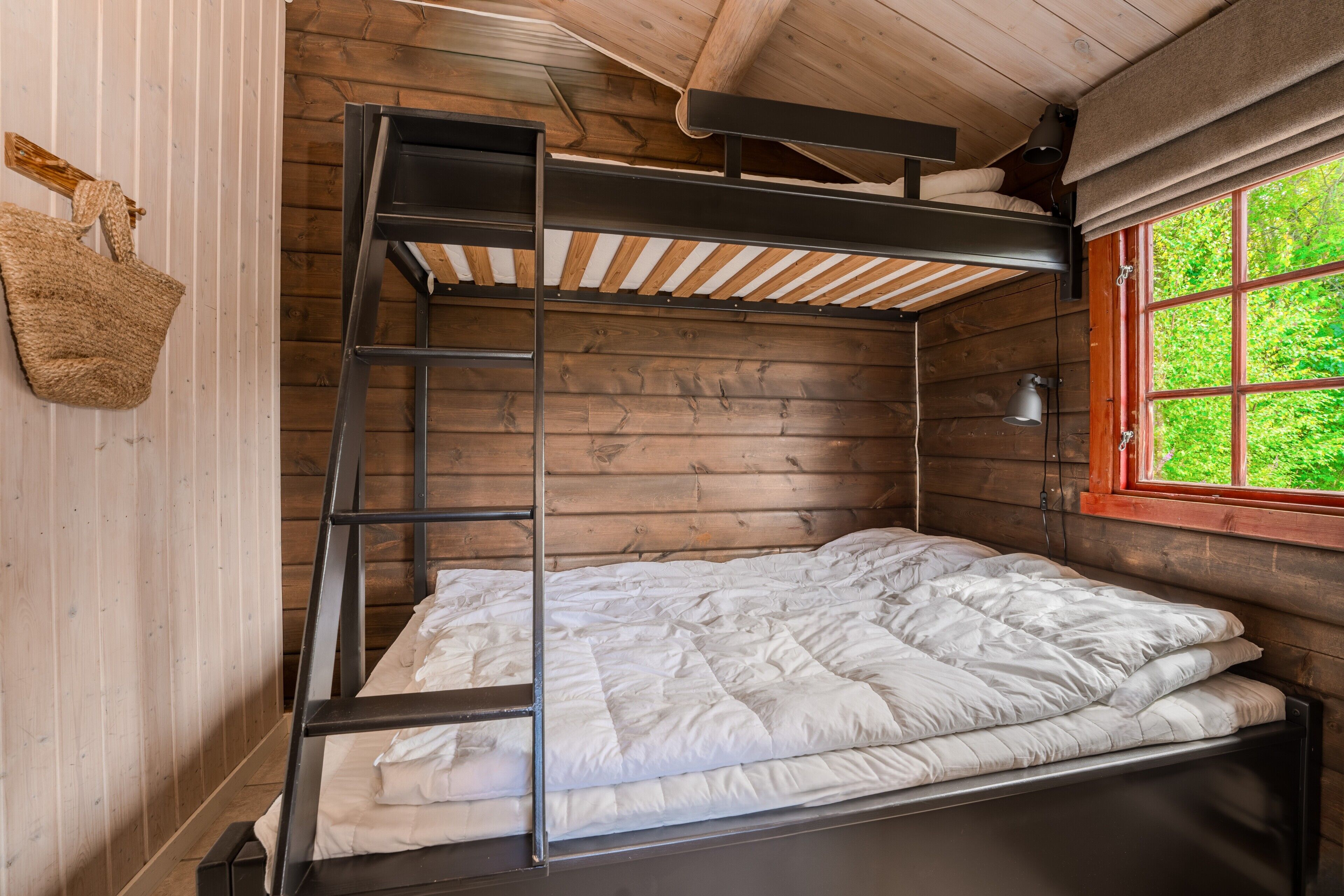 Una cama litera con escalera, una ventana con vistas a la vegetación y un bolso tejido en la pared.