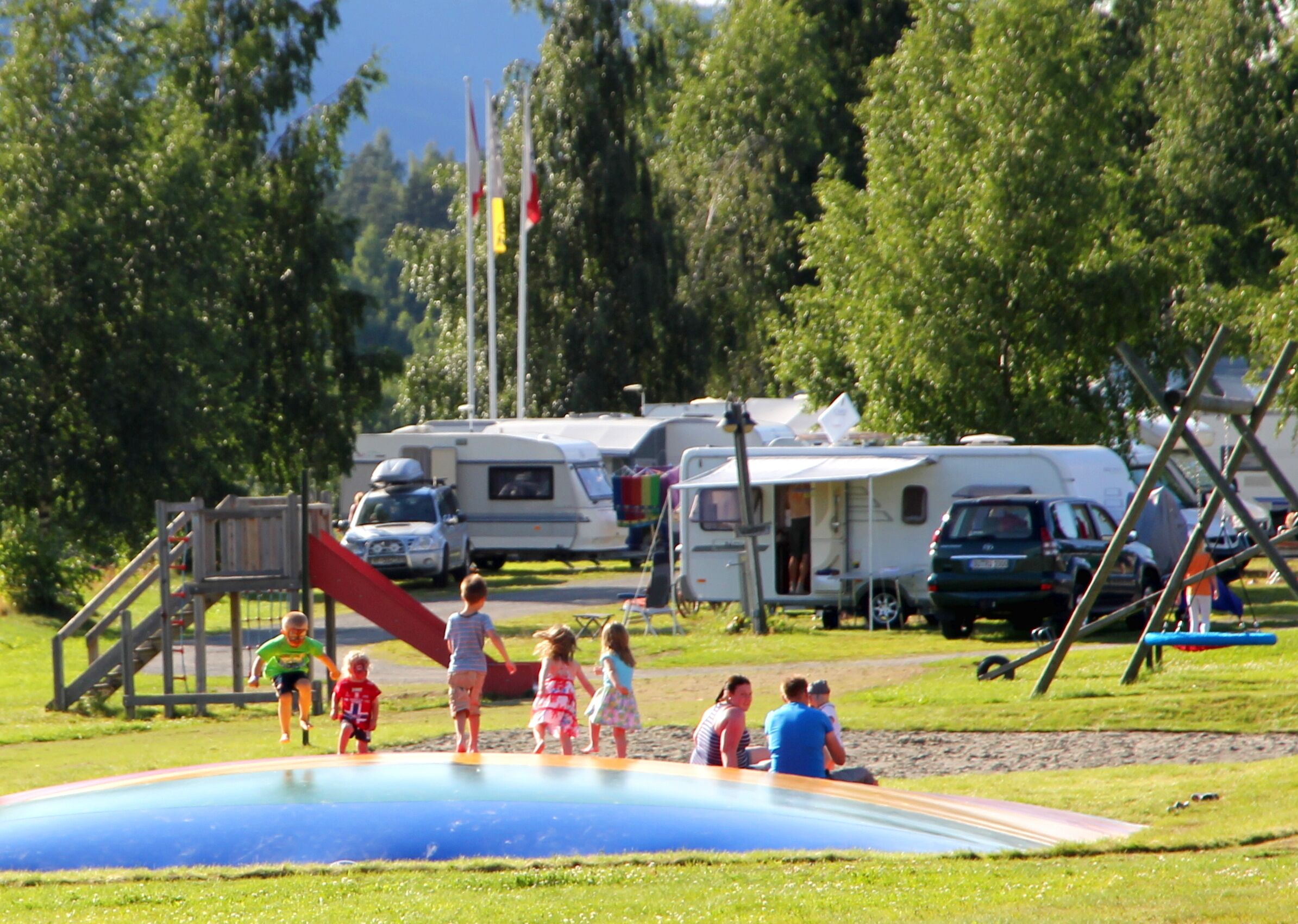 Un'area giochi con uno scivolo e una piccola piscina, circondata da alberi e camper sullo sfondo.