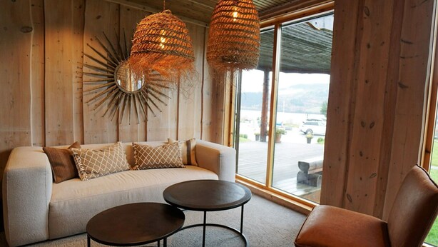 Una sala moderna con un sofá, dos mesas redondas y un sillón. Hay dos lámparas colgantes de mimbre y un adorno de pared con forma de sol.