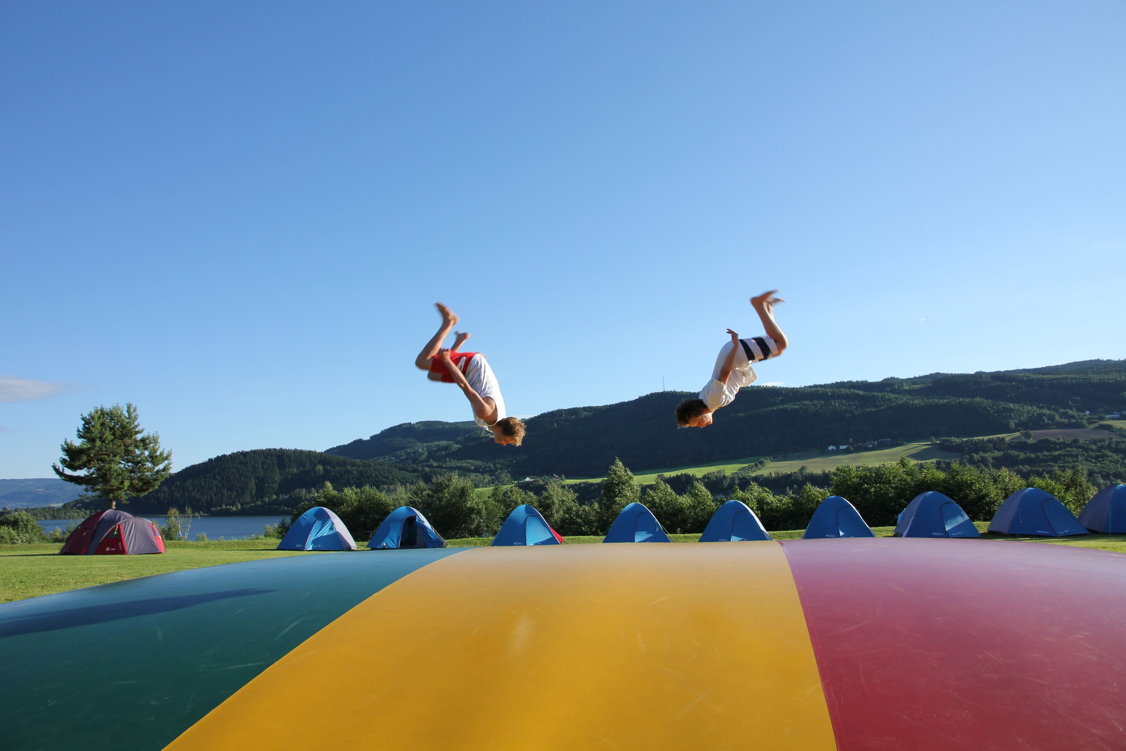 Two people mid-air performing flips on a colorful slide, with tents and a lake in the background.