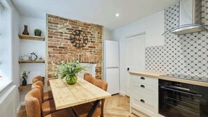 A kitchen with a wooden table, chairs, a brick wall, a clock, and a modern oven. - Host & Stay | Cragg Cottage (Whitby)