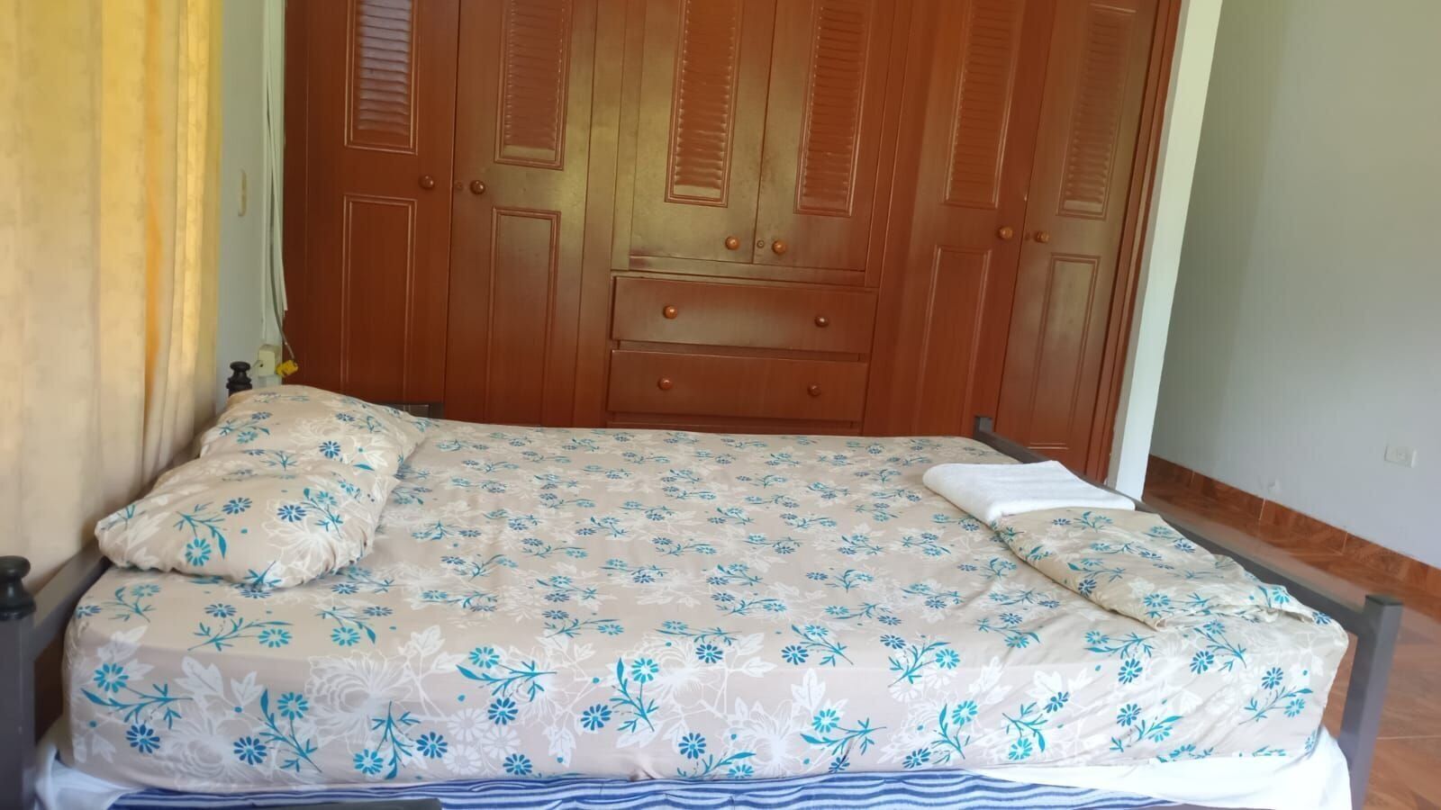 A bed with a floral bedspread, wooden wardrobe, and a curtain.