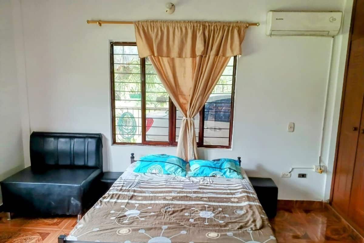 Un dormitorio con cama, sofá negro, ventana con cortinas y unidad de aire acondicionado.