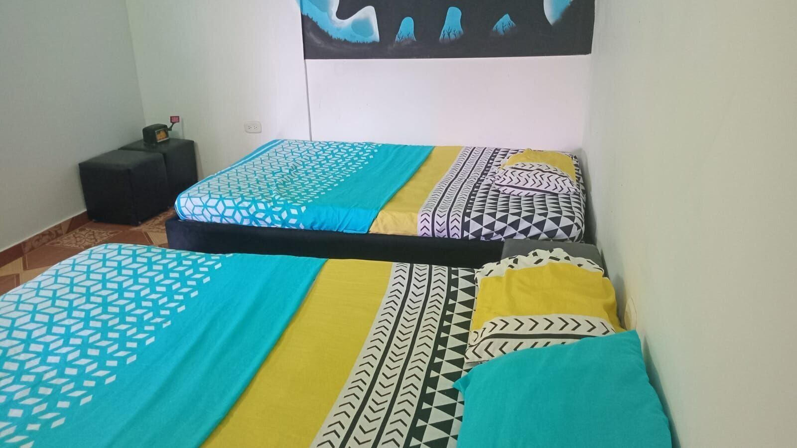 A room with two beds, each with patterned bedding. There is a small black table with a lamp beside one bed.