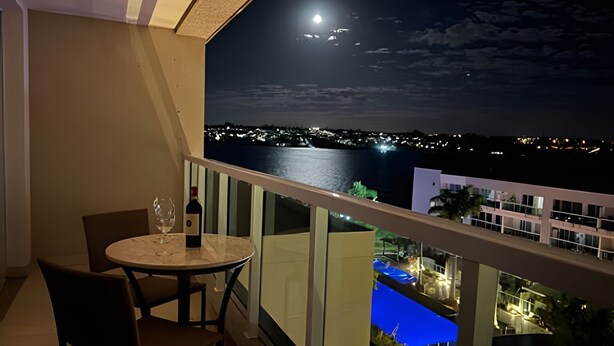 Un balcón con mesa y sillas, una botella de vino y vistas de un paisaje urbano nocturno con luna y estrellas.