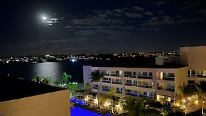 Vista nocturna de un hotel frente al mar, con zona de piscina y un paisaje urbano al fondo.