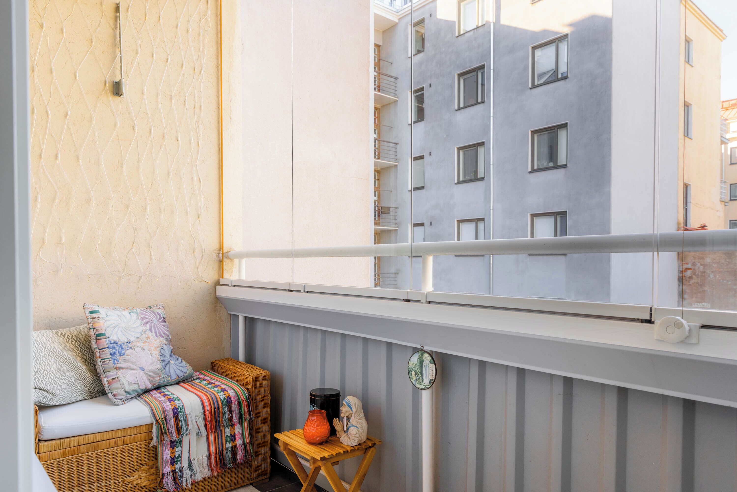 A balcony with a wicker sofa, a small wooden table with a vase and a figurine, and a view of a multi-story building.