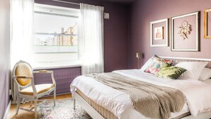 Une chambre à coucher avec un lit, une chaise, une fenêtre avec des rideaux et des tableaux encadrés accrochés au mur.